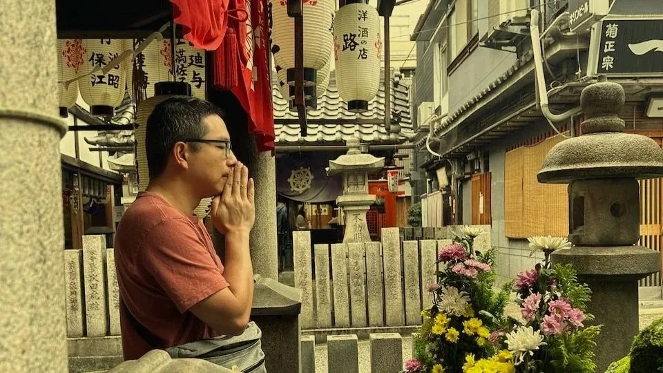 Rick, in glasses and a rust-red T-shirt, stands with his hands pressed together in prayer at a small shrine in Japan, beneath red banners and paper lanterns, with stone lanterns, carved stone posts and bright flowers around him.