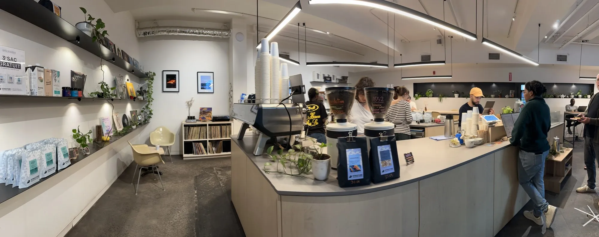 Wide view inside Structure: a long wooden coffee bar with grinders and bags of beans, baristas at work, customers ordering, and shelves of retail coffee and records along the wall.